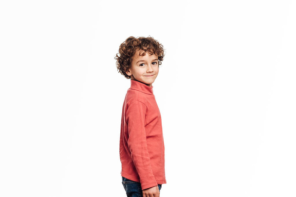 Νεαρό αγόρι κοιτάζει την κάμερα και χαμογελά ενώ στέκεται σε ένα απομονωμένο φόντο. - Φωτογραφία, εικόνα