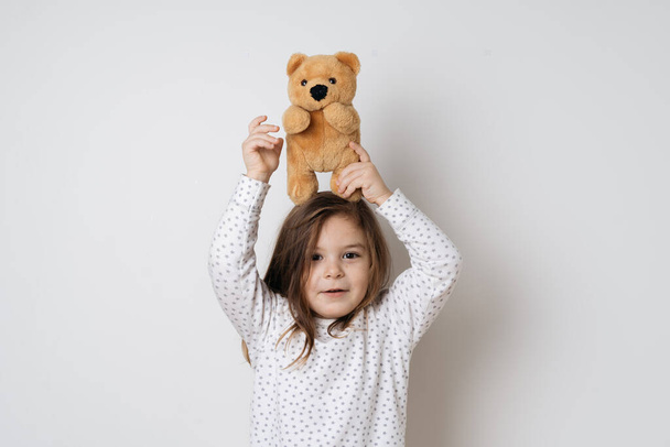 Young pretty caucasian girl with vintage teddy bear in pyjamas in white studio  - Foto, Bild