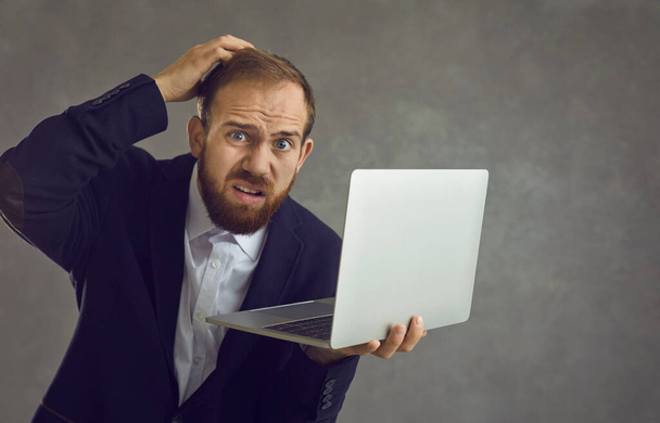 Confused businessman holding a laptop and scratching his head while standing on a gray background. - Foto, Imagen