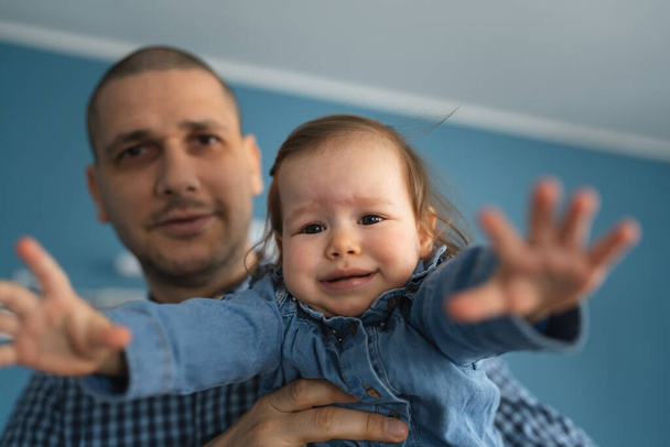 One frustrated baby trying to reach camera crying in hands of her father holding her while standing at home wearing blue shirt real people copy space - Photo, Image
