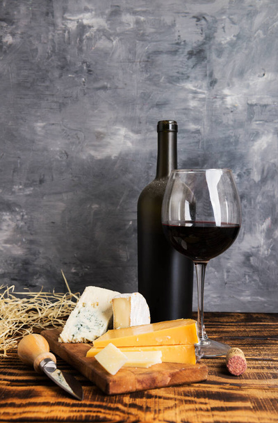 Dusty bottle with Red wine with cheese on a wooden table against a gray wall - Фото, изображение