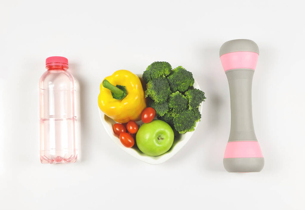 Widok z góry warzyw capsicum, brokuły, pomidory i jabłko w kształcie serca płyta na białym tle z różowej butelki wody i szary różowe hantle. Zdrowy styl życia. - Zdjęcie, obraz