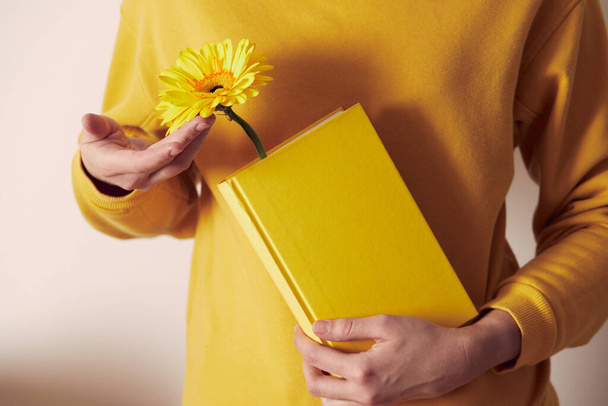 A yellow book with a flower bookmarked in the hands of a woman on a yellow sweater background. - Foto, afbeelding