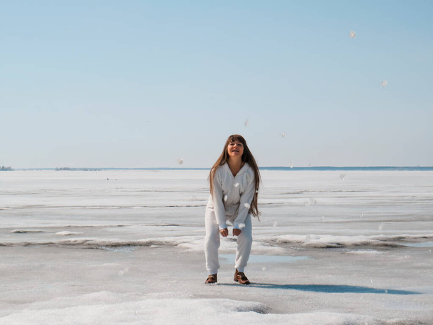 Happy beautiful woman with long dark hair in a white jumpsuit on the ice of a frozen lake on a warm spring day in Siberia, the concept of an active and healthy lifestyle, outdoor recreation, travel and hiking, seasons - Photo, image