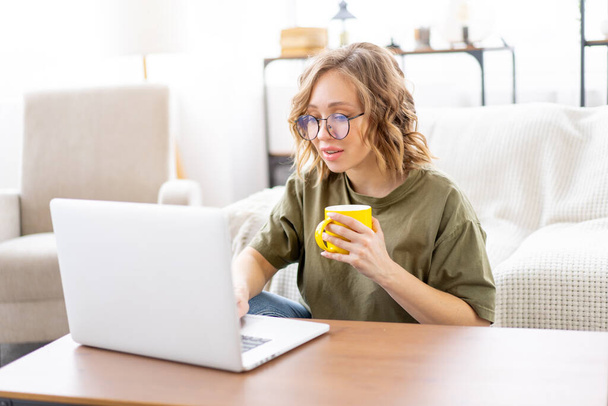 Woman with glasses use laptop drink morning coffee sitting floor near  sofa big window background home interior Freelance female working home Distance learning student relaxing watch video lesson - Foto, Imagem