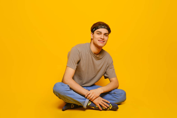 Happy young man in everyday clothes sitting on the floor in bright yellow studio - Foto, Imagen
