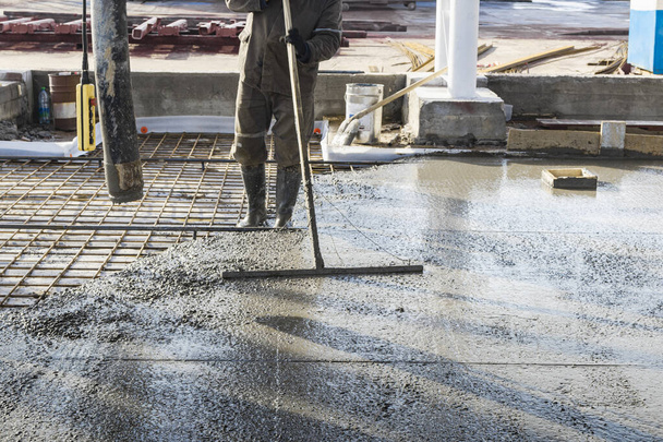 Építőipari munkás szintező öntött beton padló egy ipari műhelyben. Lábak csizmában a betonban. Felszíni betonozás. Monolitikus vasbeton gyártása - Fotó, kép