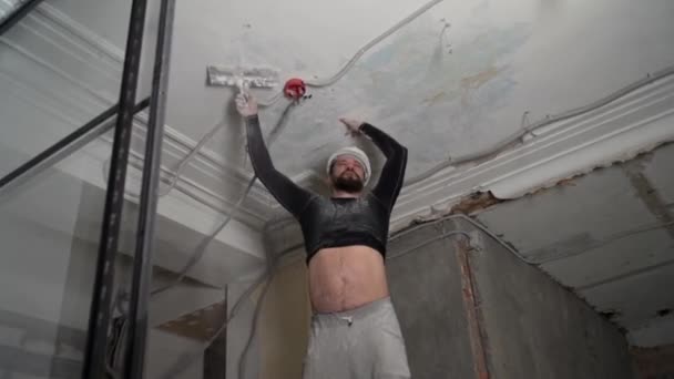 Koruyucu kasklı ve siyah dar elbiseli şişman sakallı yetişkin adam inşaat beyazlatıcısının yüksek tavanını kaldırıyor. Tamiratı kendi el konseptinle yap. 4k ağır çekim görüntüleri  - Video, Çekim