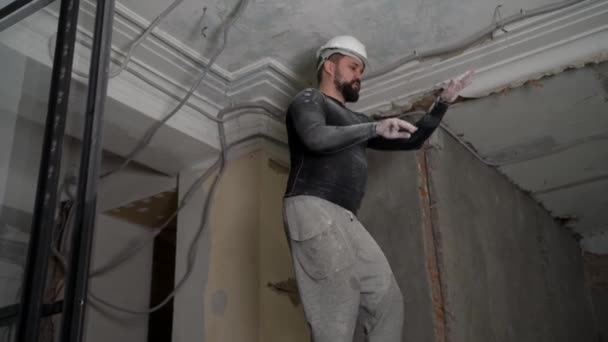 Koruyucu kasklı ve siyah dar elbiseli şişman sakallı yetişkin adam inşaat beyazlatıcısının yüksek tavanını kaldırıyor. Tamiratı kendi el konseptinle yap. 4k ağır çekim görüntüleri  - Video, Çekim