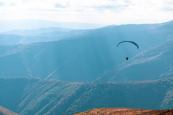 Paragliding active sports with parachute flying against mountains with lush forests in highland illuminated by sunlight under cloudy sky - Фото, зображення