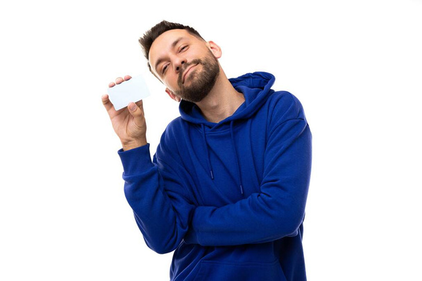 un jeune homme en sweat-shirt bleu sur fond blanc tient une carte bancaire dans ses mains - Photo, image