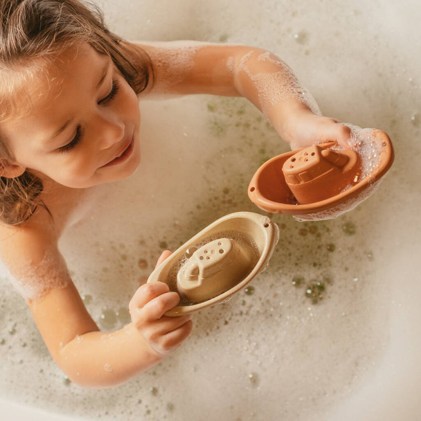 Roztomilé usmívající se holčička sedí v bublinkové koupeli a hraje si s roztomilé malé béžové a oranžové silikonové hračky lodě. - Fotografie, Obrázek