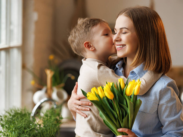 Молодая женщина мать с цветочным букетом обнимает сына, получая поздравления с Днем матери - Фото, изображение