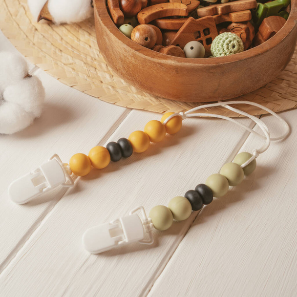 Pinza de plástico blanca con perlas de madera blanca, negra y naranja en cordones blancos para chupetes de bebé cerca de un cuenco de madera. - Foto, imagen