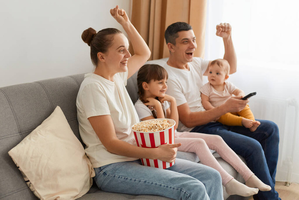Side view portrait of happy couple with their children sitting on sofa with children and watching tv or movie, rejoicing happy episode in film, screaming with raised arms and clenched fists. - Фото, изображение