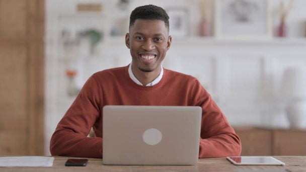Homem Africano sorrindo para a câmera ao usar o laptop no escritório - Foto, Imagem