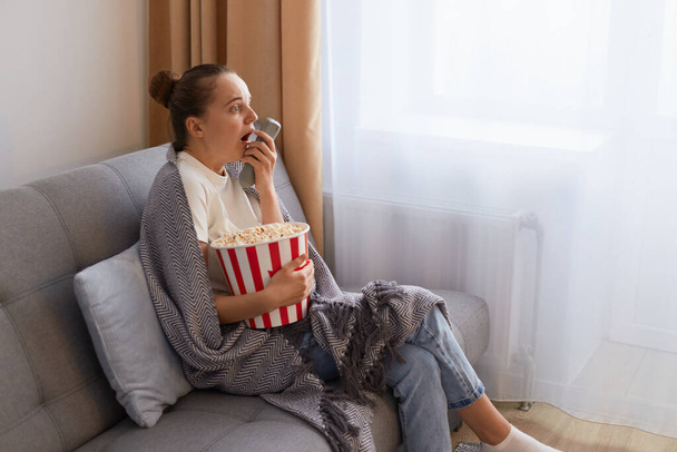 Profile portrait of of attractive socked scared woman with bun hairstyle sitting on couch wrapped in blanket and watching scary movie, embracing bucket with popcorn, keeps mouth open. - 写真・画像