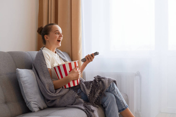 Profile portrait of of attractive impressed woman with bun hairstyle sitting on couch wrapped in blanket and watching film with tasty snack, enjoying interesting film or tv show. - 写真・画像