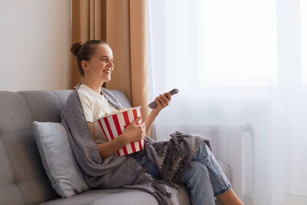 Side view of woman with bun hairstyle sitting on sofa wrapped in gray blanket and watching movie and eating popcorn, enjoying interesting film or tv show. - 写真・画像
