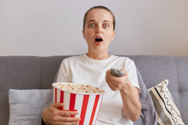 Portrait of shocked woman wearing white casual style t shirt, sitting on couch and holding paper bucket with popcorn, watching horror film alone, keeps mouth widely open. - Foto, immagini