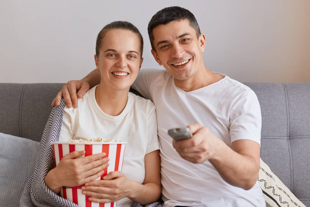 Indoor shot of attractive young couple watching films together while sitting on sofa at home, man hugging her wife, family expressing positive emotion, love and care. - Fotó, kép