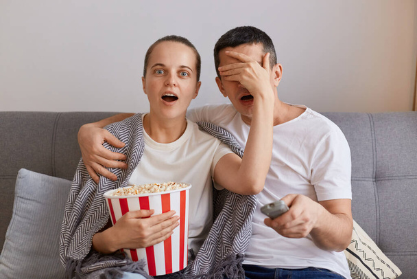 Portrait of impressed woman watching horror film with man, shocked wife covered husband's eyes with palm, forbidden content on tv, couple sitting on sofa wrapped in blanket. - Photo, Image