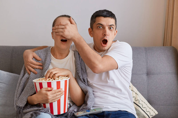 Image of impressed man watching horror film with woman, shocked husband covered his wife's eyes with palm, forbidden content on tv, couple sitting on sofa wrapped in plaid - Photo, image
