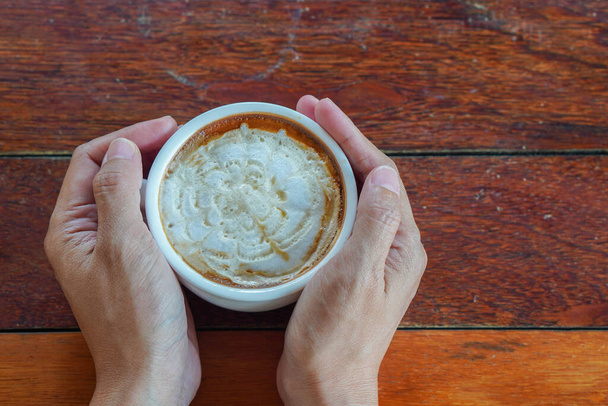 Foto links, bovenaanzicht, twee handen met een hete karamel macchiato koffiekop op een houten vloer achtergrond, eten, drinken, kopieer ruimte - Foto, afbeelding