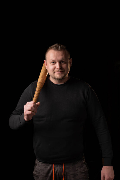 A young man is holding a wooden bat. Bouncer, security guard, bodyguard concept. A large man in a T-shirt looks at the camera. - Foto, afbeelding