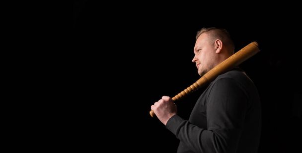 Young man with a baseball bat on a dark background. Bouncer, security guard, bodyguard concept. Place for text. A large man in a T-shirt stands sideways. - Φωτογραφία, εικόνα