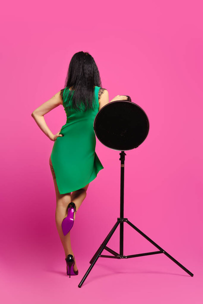 Rear view of woman with black hair and in green dress rising one leg and leaning on studio strobe - Photo, image