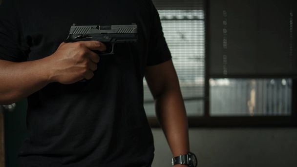 Мужчина, держащий пистолет, стоящий в комнате в черном, указывающий и нацеленный на цель. Понятие преступления, убийства, криминала - Фото, изображение