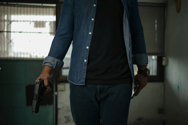 A man in a blue jeans shirt with a mask and a silver cap is standing in the room with a pistol. Point and aim the gun at the target. concept of assassination, murder, criminal - Photo, Image