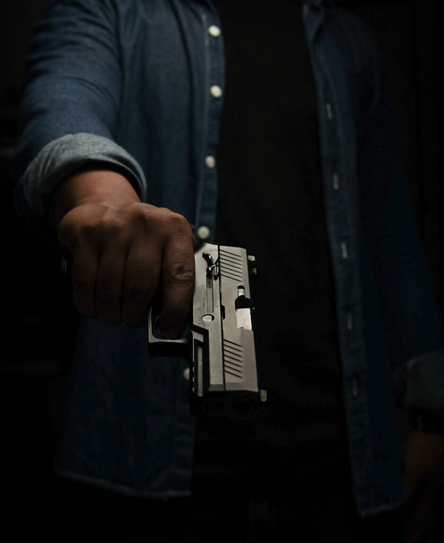 A man in a blue denim shirt is standing in a dark room with a pistol. Point and aim the gun at the target. concept of assassination, murder, criminal - Photo, image
