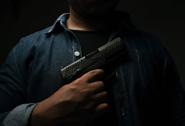 Mężczyzna w niebieskiej koszuli stoi w ciemnym pokoju z pistoletem przystawionym do piersi. koncepcja zabójstwa, morderstwa, przestępstwa - Zdjęcie, obraz