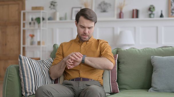 Jeune homme attendant tout en vérifiant la montre sur le canapé - Photo, image
