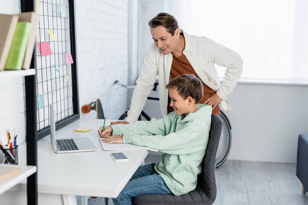 Père souriant debout près de fils écrit sur ordinateur portable près des appareils à la maison  - Photo, image