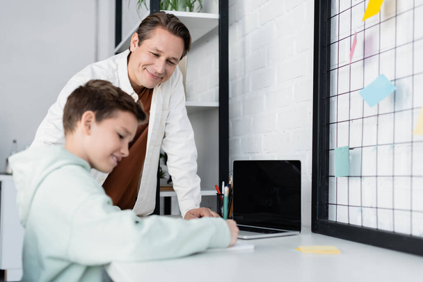 Positive man looking at blurred son writing on notebook near laptop with blank screen at home  - Photo, Image