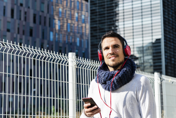 Atractivo joven escuchando música por auriculares mientras sostiene un teléfono móvil al aire libre - Foto, imagen