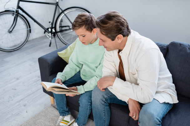 Smiling man sitting near son reading book on sofa in living room  - Photo, Image