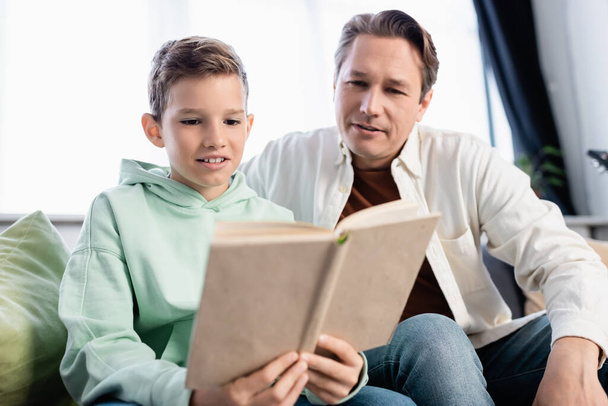 Nastolatek i ojciec czytający zamazaną książkę w salonie  - Zdjęcie, obraz