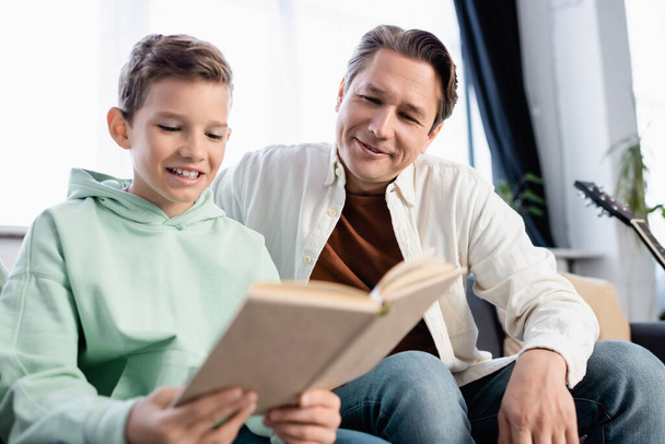 Garçon souriant lecture livre flou près de papa à la maison  - Photo, image