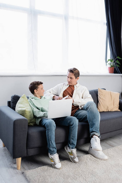 Smiling child using laptop near dad with cellphone on couch  - Photo, image