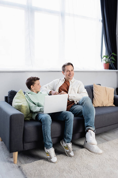 Homem positivo segurando smartphone perto do filho com laptop na sala de estar  - Foto, Imagem