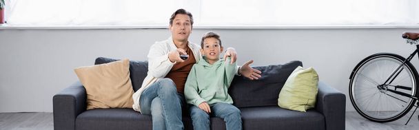 Excited man with remote controller watching movie with son at home, banner  - Фото, изображение