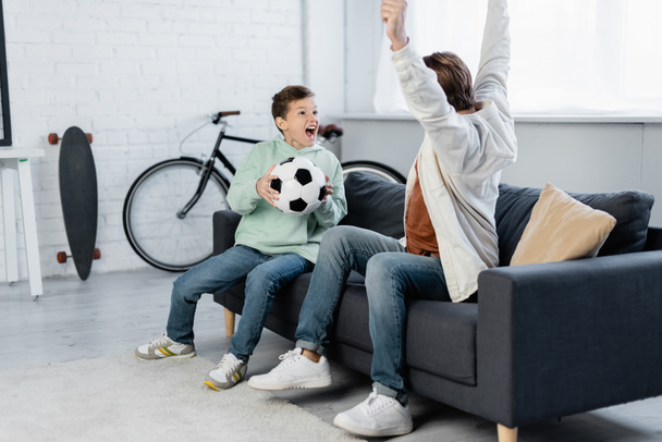 Aufgeregtes Vorschulkind hält Fußballball neben Papa auf der heimischen Couch  - Foto, Bild