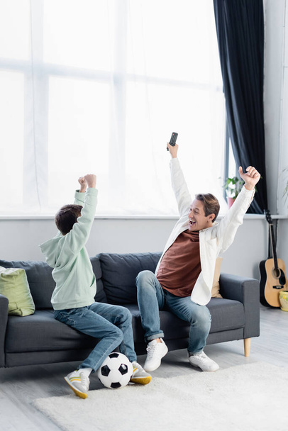 Emocionado padre sosteniendo mando a distancia y mostrando sí gesto cerca de hijo y pelota de fútbol en casa  - Foto, Imagen