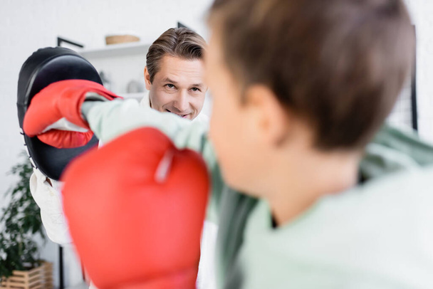 Pozitivní muž trénink s rozmazané předpubertální syn v boxerských rukavicích doma  - Fotografie, Obrázek