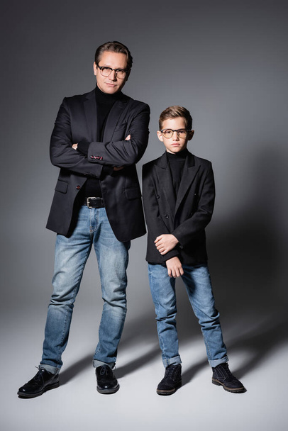 Full length of stylish boy in jacket and jeans standing near father on grey  - Zdjęcie, obraz