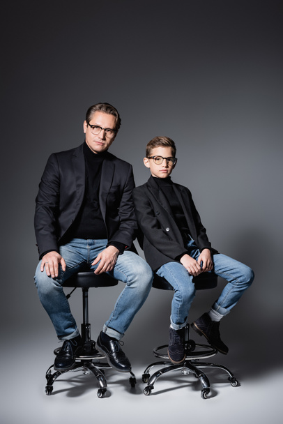 Stylish father and preteen son sitting on chairs on grey  - 写真・画像